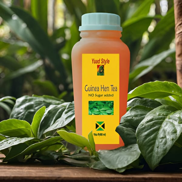 A bottle of Yaad Style Guinea Hen Tea, with a Jamaican flag, on a wooden surface surrounded by green leaves.